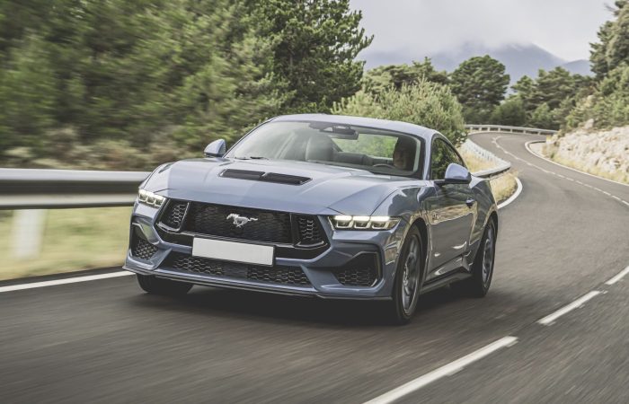 Blauer Ford Mustang GT fährt auf einer kurvenreichen Landstraße, umgeben von Bäumen und Bergen im Hintergrund